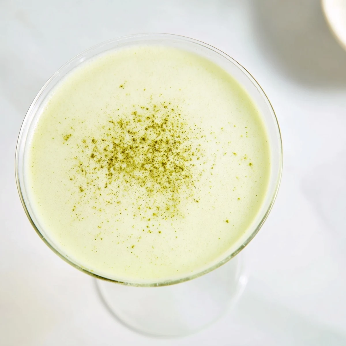 Frothy and vibrant, a Matcha Martini Cocktail sits on a marble counter, garnished with white chocolate shavings for a sweet finish.