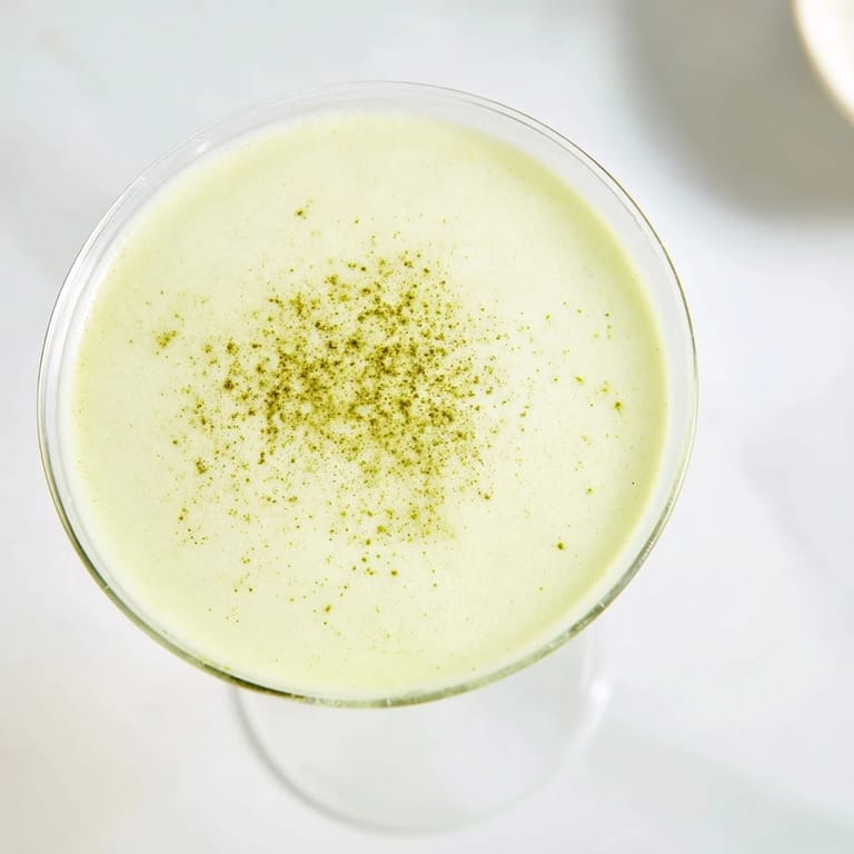 Frothy and vibrant, a Matcha Martini Cocktail sits on a marble counter, garnished with white chocolate shavings for a sweet finish.